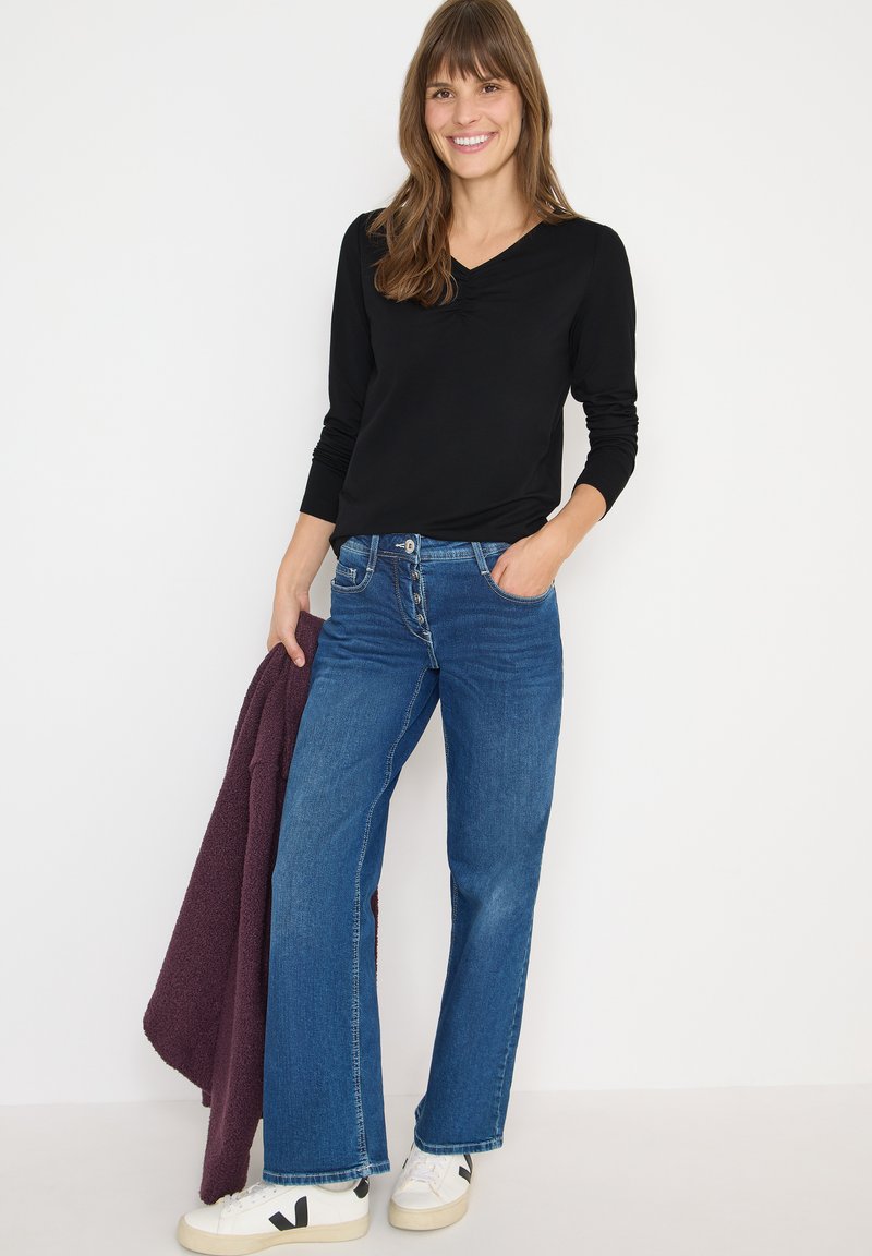 Maglietta nera a maniche lunghe, jeans blu a gamba larga, sneakers bianche con dettagli neri e un cardigan a maglia bordeaux appoggiato su un braccio.