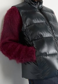 Person som bär en blänkande svart pufferväst över en fluffig mörkröd tröja, med handen i västfickan, visad från halsen till höfterna.