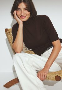 Brown ribbed turtleneck top paired with wide white pants, seated on a woven chair, featuring smooth skin and relaxed posture.