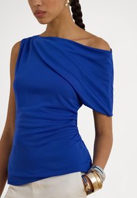 Ženska, oblečena v modro asimetrično enoročno majico, bež hlače, več zlatih in modrih zapestnic ter z ravno speto pričesko.