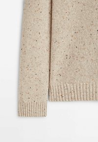 Beige neulepaita, jossa on kirjavapinta oransseilla ja mustilla raitoineen, röyhelösuukappaleet ja helma sekä pehmeä, lämmin viimeistely.