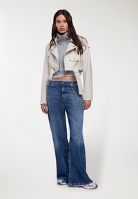 Giacca corta in pelle bianca sopra un dolcevita grigio, abbinata a jeans blu a gamba larga con dettaglio catena. Design semplice e moderno.