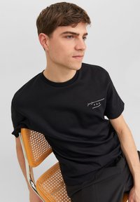 Camiseta de algodón negra con cuello redondo, mangas cortas y un pequeño logotipo blanco en el pecho. Modelo sentado en una silla con asiento de mimbre.