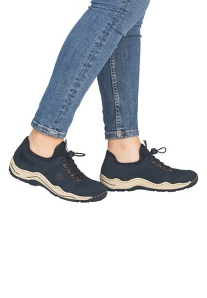 Person wearing dark blue slip-on sneakers with beige soles and orange lace details, paired with fitted blue jeans.