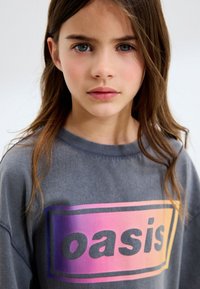 Niña joven con cabello largo y castaño, vestida con una sudadera gris que tiene un logotipo "oasis" con un degradado colorido en el frente, mirando directamente hacia adelante.