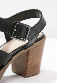 Sandale en cuir noir avec une boucle argentée et un talon épais en bois empilé, vue en gros plan.