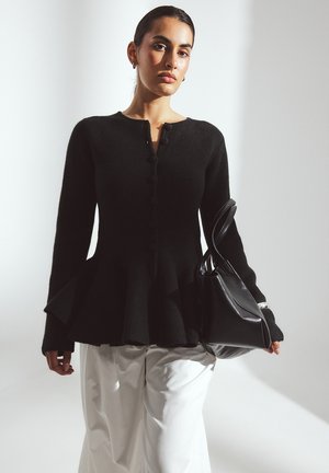 Black wool peplum cardigan with button front, long sleeves, and textured fabric; paired with white trousers and a black handbag.