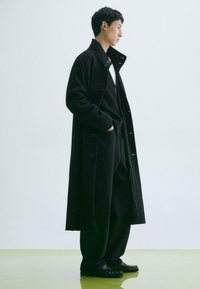 Lungo cappotto nero realizzato in un tessuto morbido, caratterizzato da un colletto alto e chiusura con bottoni, indossato sopra un maglione nero strutturato e pantaloni larghi.