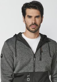 Sudadera con cierre en gris y negro, con capucha ajustable, texturas en contraste, bolsillos frontales con cremallera y acentos de logo en la manga.