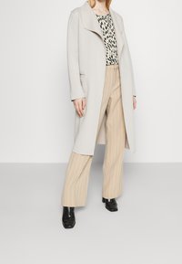 Cappotto lungo grigio chiaro con colletto a scialle, abbinato a un top a scacchi bianco e nero e pantaloni beige a righe. Stivali alla caviglia neri lucidi.