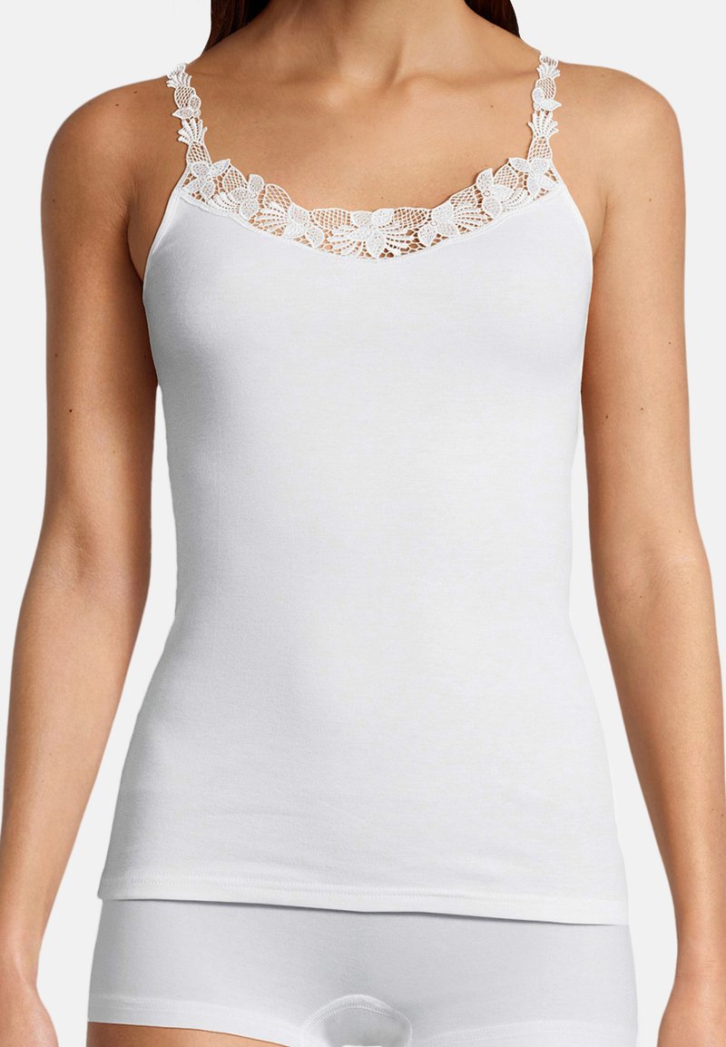 Kobieta ubrana w białą dopasowaną koszulkę na cienkich ramiączkach z kwiatową koronką na ramiączkach i dekolcie.