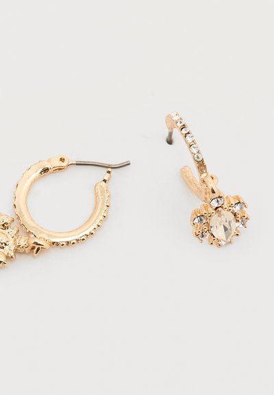 Σκουλαρίκια κρίκοι σε χρυσό τόνο με διάφανα στρας, το ένα με πέτρα σε σχήμα σταγόνας περιτριγυρισμένη από μικρότερα στρας σε λευκό φόντο.