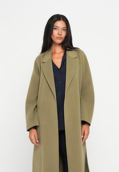 Lunga giacca verde oliva con una vestibilità su misura, ampie revers, e tasche laterali, indossata sopra una camicia button-up blu scuro. Tessuto dalla consistenza morbida.