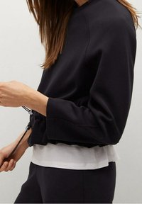 Sudadera negra con dobladillo fruncido, superpuesta a una camisa blanca. Presenta un escote redondo y mangas largas con puños anchos.