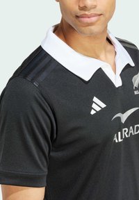 Sort rugbyshirt med hvid krave, der har tre mørke striber på skuldrene og sølvlogo-accenter. Stoffet er tekstureret og tætsiddende.