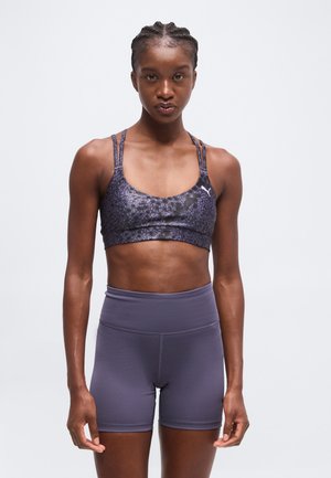 Femme athlétique aux cheveux tressés porte un soutien-gorge de sport sombre à motifs et un short de sport violet taille haute, debout devant un fond uni.