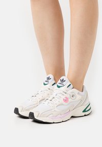 Vita läder- och nät-sneakers med gröna och rosa accenter, texturerade detaljer och en svart gummisula. Adidas-logotyp på tungan.