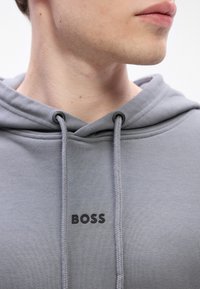 Detailný záber dolnej časti tváre a krku osoby, ktorá má na sebe svetlošedú mikinu s kapucňou, na hrudi s čiernymi písmenami slovo "BOSS".