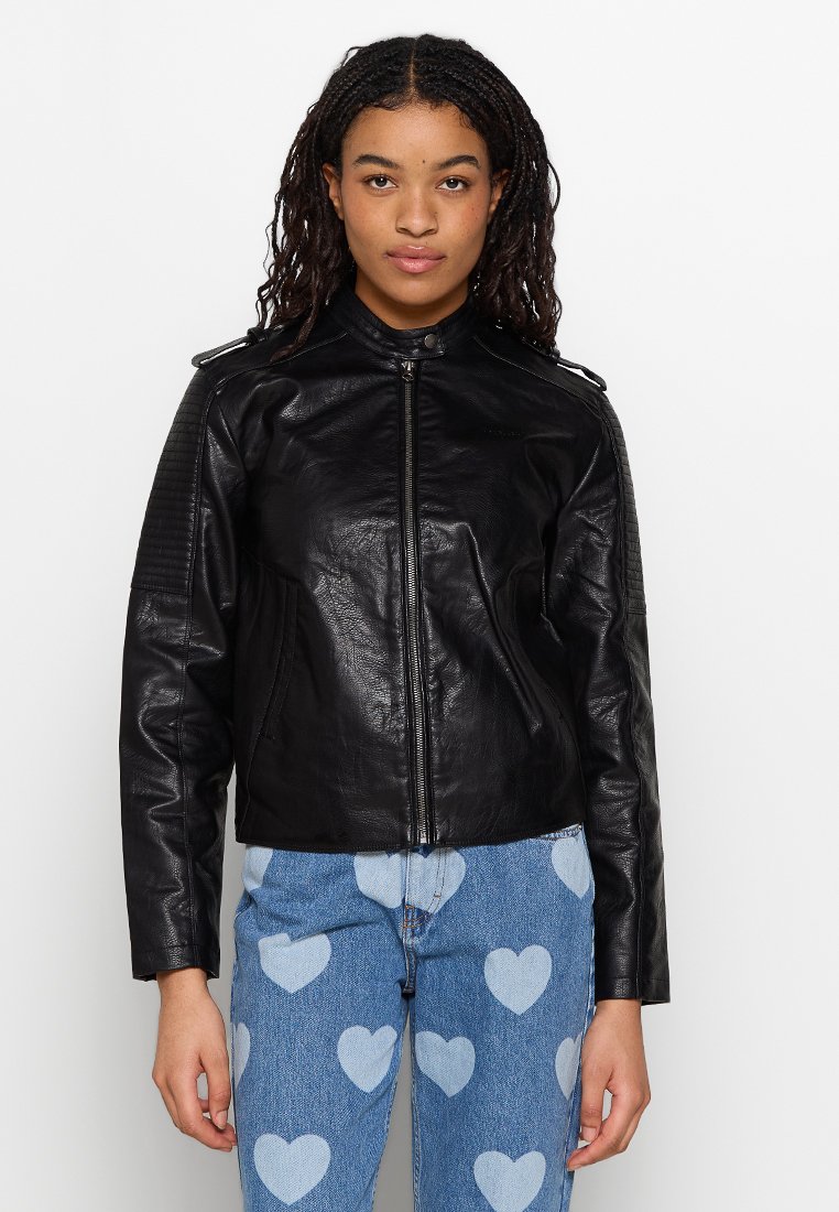 Chaqueta de motociclista de cuero negro con cierre frontal, cuello alto y detalles de hombros acolchados, combinada con jeans azules con patrones de corazones.