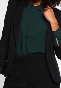 Zwarte geribde blazer over een donkergroene blouse met kraag, met een elastische tailleband op zwarte broeken. Glad materiaal met subtiele details.