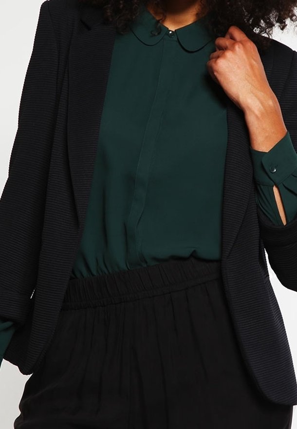 Zwarte geribde blazer over een donkergroene blouse met kraag, met een elastische tailleband op zwarte broeken. Glad materiaal met subtiele details.