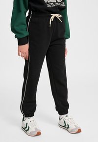 Czarne dresy z elastycznym paskiem i sznurkiem, z kontrastującymi białymi paskami po bokach i ściągaczami na dolnych nogawkach. Noszone z sportowymi butami.