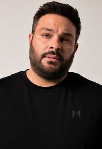 Muž s krátkými tmavými vlasy a vousy, oblečený v černém tričku s kulatým výstřihem a jemným logem "M" na hrudi, na jednobarevném pozadí.