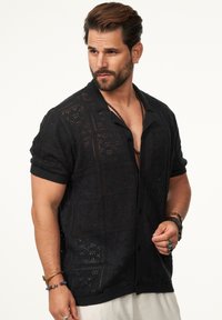Schwarzes gestricktes kurzärmeliges Hemd mit gemusterten Löchern, einem Kragen und Knöpfen. Verfügt über gerippte Kanten und eine lockere Passform.