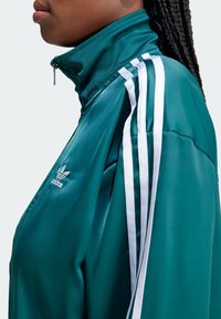 Teal urheilujakko, jossa on korkea kaulus, valkoiset raidat harteilla ja Adidas-logo vasemmalla puolella. Silkkinen, kiiltävä kangas ja istuva leikkaus.