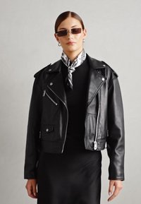 Black leather biker jacket with silver hardware, zippered pockets, and a cropped fit. Paired with a black top and a patterned scarf.