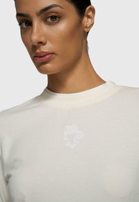 Camiseta de algodón beige con escote redondo y un sutil gráfico bordado en blanco en el pecho. Textura suave con un corte ajustado.