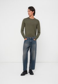 Camisa de manga larga verde oliva combinada con jeans de mezclilla azul holgados. El modelo está de pie con las manos en los bolsillos, llevando zapatos negros. Fondo liso.
