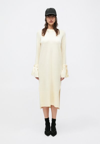 Long, cream-colored knit dress with ribbed hem and wide sleeves tied at the wrists. Paired with black cap and ankle-length black socks.