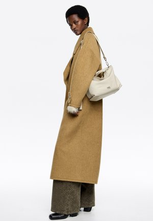 Długi, brązowy płaszcz z wełny z bocznymi kieszeniami, ozdobiony futrzanymi akcentami na rękawach; zestawiony z szerokimi, teksturowanymi oliwkowymi spodniami i jasnobeżową torebką.