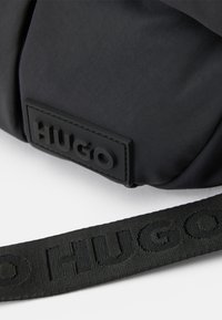 Gros plan sur un sac en tissu noir avec un patch en caoutchouc et une sangle, tous deux embossés du nom de la marque "HUGO".