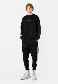 Sudadera negra con una etiqueta rectangular y pantalones jogger negros con un diseño gráfico. Llevado con botas blancas de cordones.