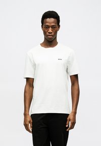 Άνδρας με κοντά σγουρά μαλλιά, φορώντας απλό λευκό μπλουζάκι με το λογότυπο "BOSS" στο στήθος και μαύρο παντελόνι, στέκεται μπροστά σε λευκό φόντο.