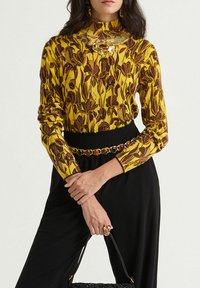 Maglia a maniche lunghe gialla con motivo floreale, abbinata a un fondo nero con cintura. Accenti dorati negli accessori e una borsa testurizzata.