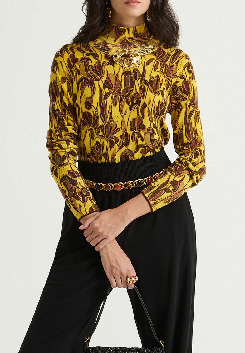 Maglia a maniche lunghe gialla con motivo floreale, abbinata a un fondo nero con cintura. Accenti dorati negli accessori e una borsa testurizzata.