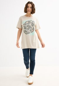 Camiseta de algodón beige con cuello redondo, mangas cortas y un gráfico floral en gris y menta, combinada con jeans ajustados de color azul oscuro y zapatillas blancas.