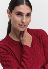 Röd texturerad tröja med diamantmönster, ribbad halsringning och långa ärmar. Modellen lutar sin haka mot en neutral bakgrund.