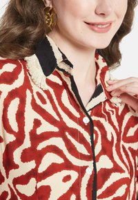 Camicia a righe rossa e crema con finiture nere, caratterizzata da un colletto testurizzato e bottoni, raffigurante un design astratto ondulato.