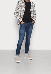 Rutig skjorta i beige och blått, svart tröja, blå jeans och ljusgrå sneakers. Lagerdesign med knäppning och avslappnad passform.