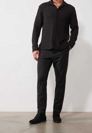 Chemise noire texturée à manches longues avec des boutons, associée à un pantalon sombre et des chaussures en daim noires. Design simple et ajusté avec un col.