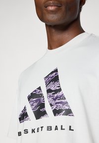 Hvid T-shirt med et sort og lilla grafisk logo, der viser tre trekantede former og teksten "BASKET BALL" i sort. Fremstillet af bomuld.