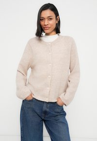 Ανοιχτό μπεζ ζακέτα από απαλό υλικό, με στρογγυλή λαιμόκοψη, μπροστινά κουμπιά και μακριά μανίκια. Φορεμένη πάνω από λευκή μπλούζα και συνδυασμένη με μπλε τζιν.