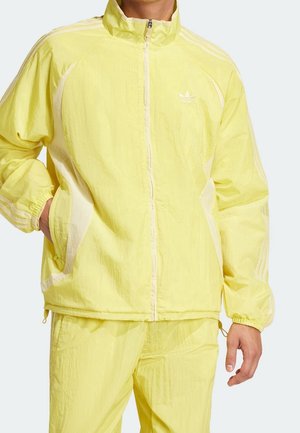 Mężczyzna ubrany w jasny żółty dres Adidas z białymi paskami, kurtkę na zamek i dopasowane spodnie, stojący z jedną ręką w kieszeni.