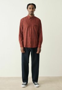 Punane ruuduline nööpidega särgiga, millel on rinnatasku; kantud tumehallide sirgete pükstega ja mustade tennistega, neutraalse tausta ees.