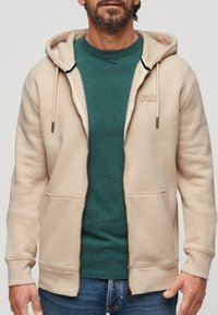 Sudadera beige con cremallera y cordones, bolsillos frontales y un logo sutil. Llevada sobre una camiseta de cuello redondo de color teal y jeans azules. Textura suave.