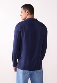 Marineblaues Langarmshirt mit lässiger Passform. Weiche Textur, einzelner Knopfverschluss am Ärmel und Rückenpasse-Detail. Kombiniert mit hellblauen Jeans.